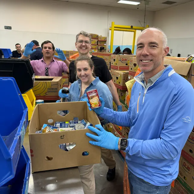 Community Food Collection Effort A group of volunteers smiling while sorting food items in a warehouse. One person is holding a can, and several boxes are visible in the background.