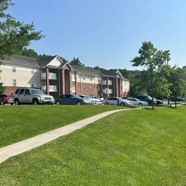 A view of a residential apartment complex with cars parked in front, surrounded by green grass and trees under a clear blue sky.
