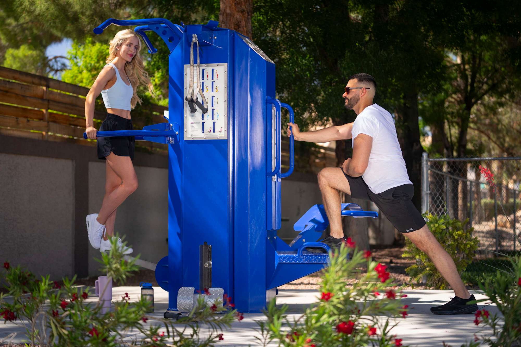 Two individuals exercising on an outdoor gym equipment. One person is seated, using a machine while the other is standing beside it, both smiling.
