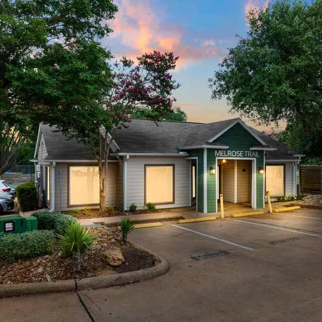 Exterior view of a small building named Melrose Trail, surrounded by trees and greenery with a sunset sky in the background.