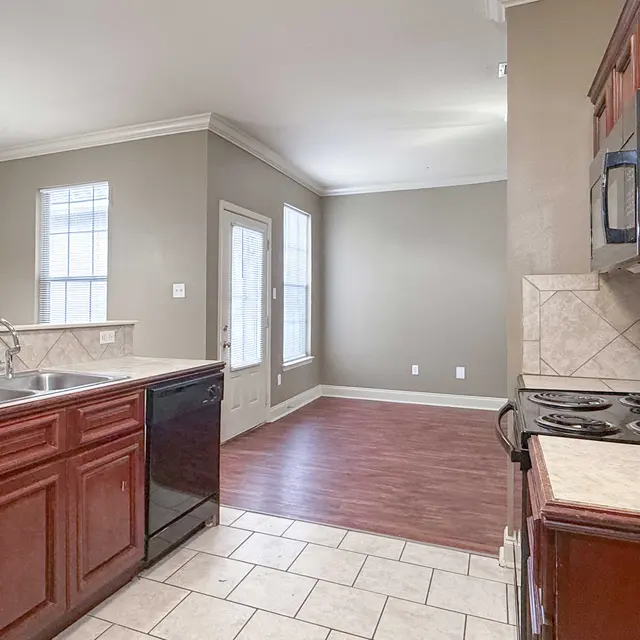 A modern kitchen featuring dark wood cabinets, a black refrigerator, and a stainless steel stove with a tiled backsplash. There's a tile floor and a view into a living space with wood laminate flooring and large windows.