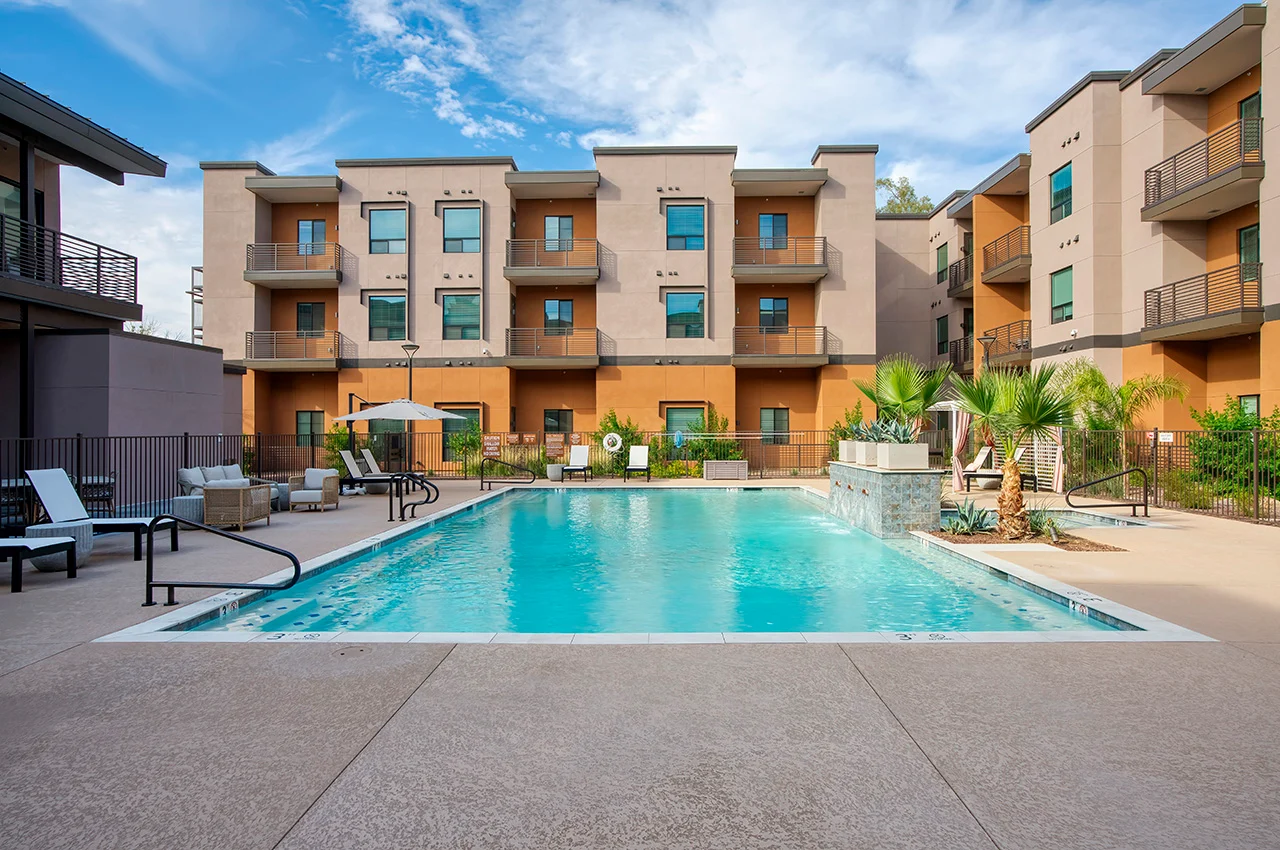 A swimming pool surrounded by lounge chairs and modern apartment buildings, with palm trees and a clear blue sky.