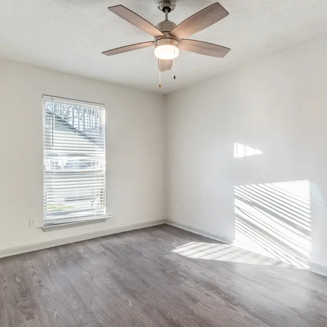 A bright, empty room featuring a ceiling fan, a window with blinds, and light wooden flooring.