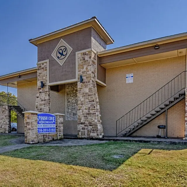 A two-story building with a stone facade and a sign for Spanish Cove Apartments. The building features a staircase on the right side and a grassy area in front.