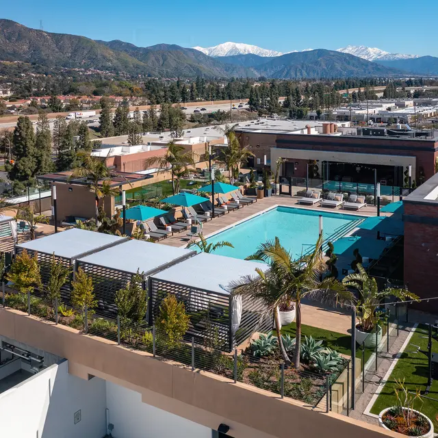 Aerial view of a modern rooftop pool area surrounded by lounge chairs and palm trees, with mountains in the background and clear blue skies.