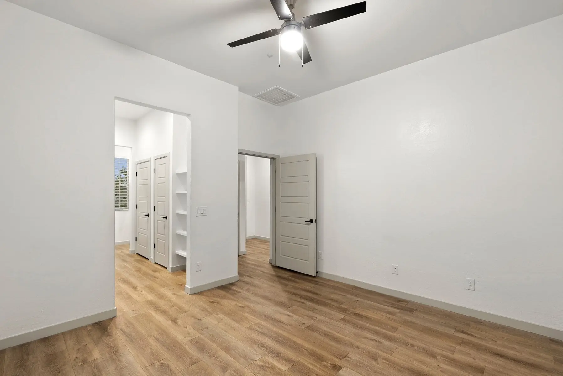 A bright, empty room with light wood flooring, a ceiling fan, and two doors leading to adjacent spaces.