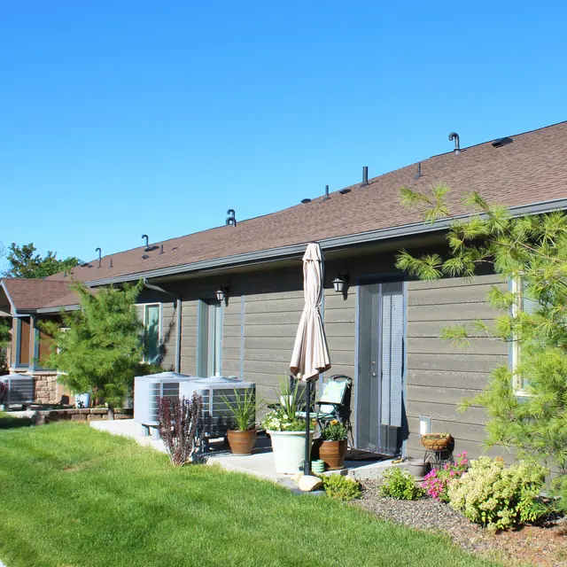 A row of single-story houses with brown siding and sloped roofs under a clear blue sky. Each house has a patio area with outdoor furniture and potted plants, surrounded by green grass.