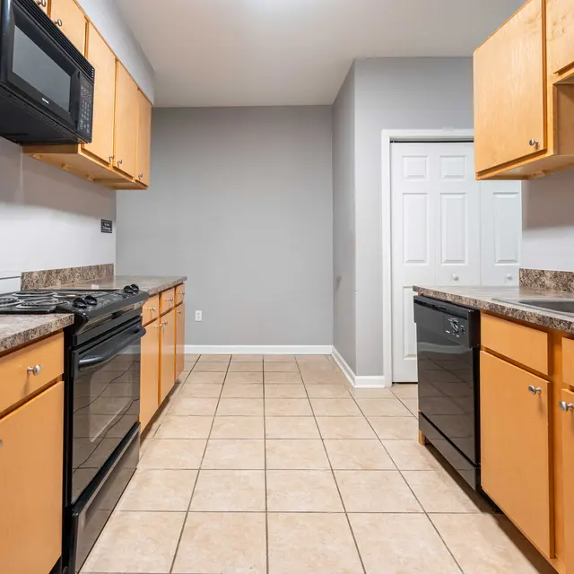 A modern kitchen featuring wooden cabinets and black appliances. The floor is tiled with a light-colored grout, and there is a door visible leading to another area.