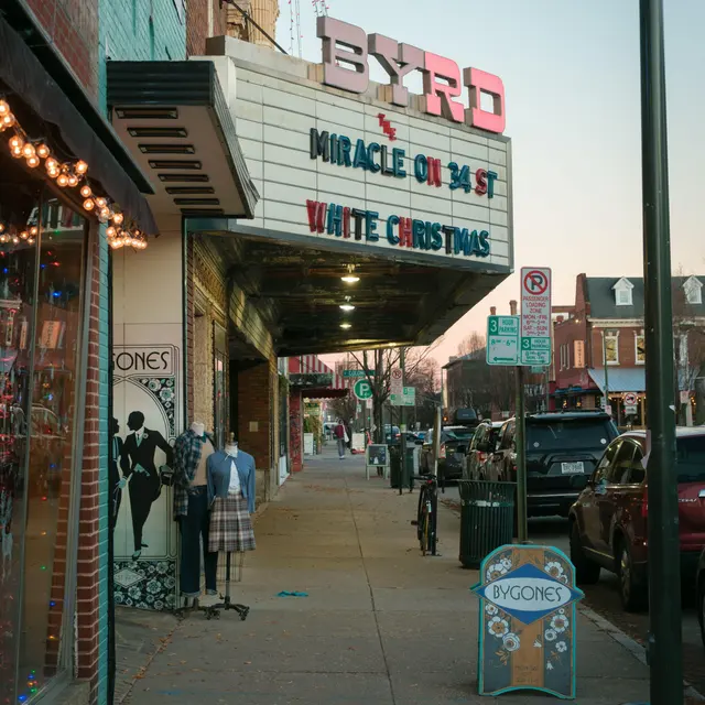 The exterior of the Byrd Theatre in Carytown, Richmond, VA featuring a marquee displaying movie titles 'Miracle on 34th St' and 'White Christmas', with a sidewalk view showing boutique shops and parked cars.