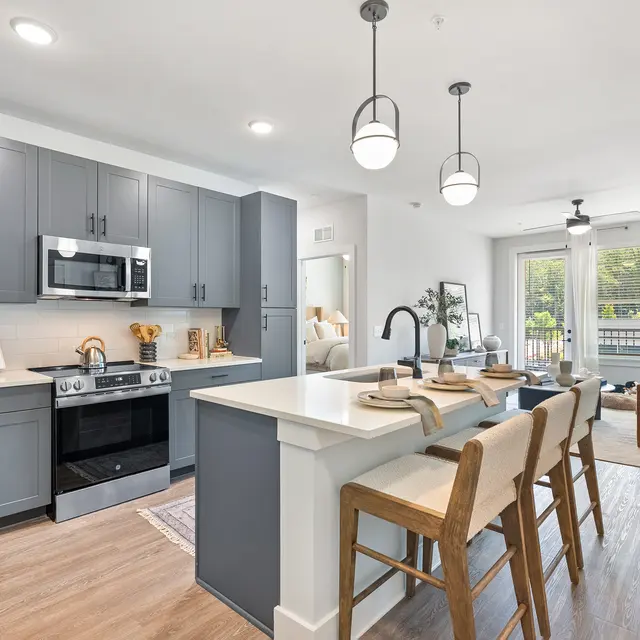 A modern kitchen with gray cabinets, stainless steel appliances, a large island with seating, and a living area featuring a brown sofa and natural light from large windows.