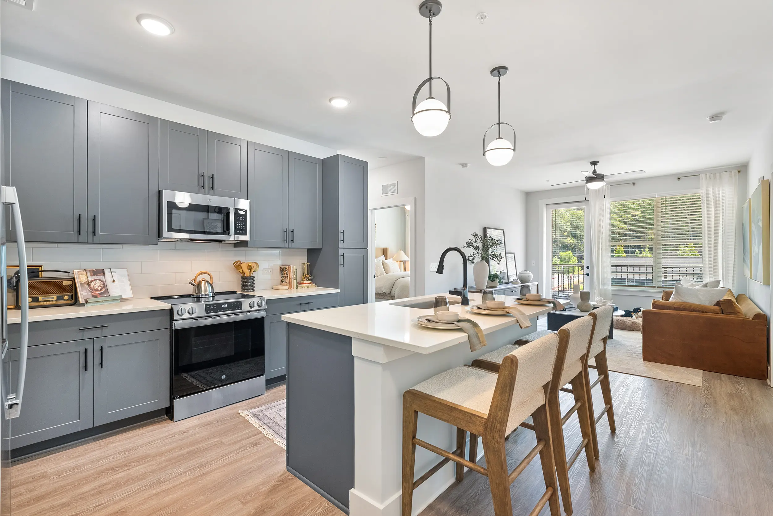 Stylish Modern Kitchen and Cozy Living Space A modern kitchen with gray cabinets, stainless steel appliances, a large island with seating, and a living area featuring a brown sofa and natural light from large windows.