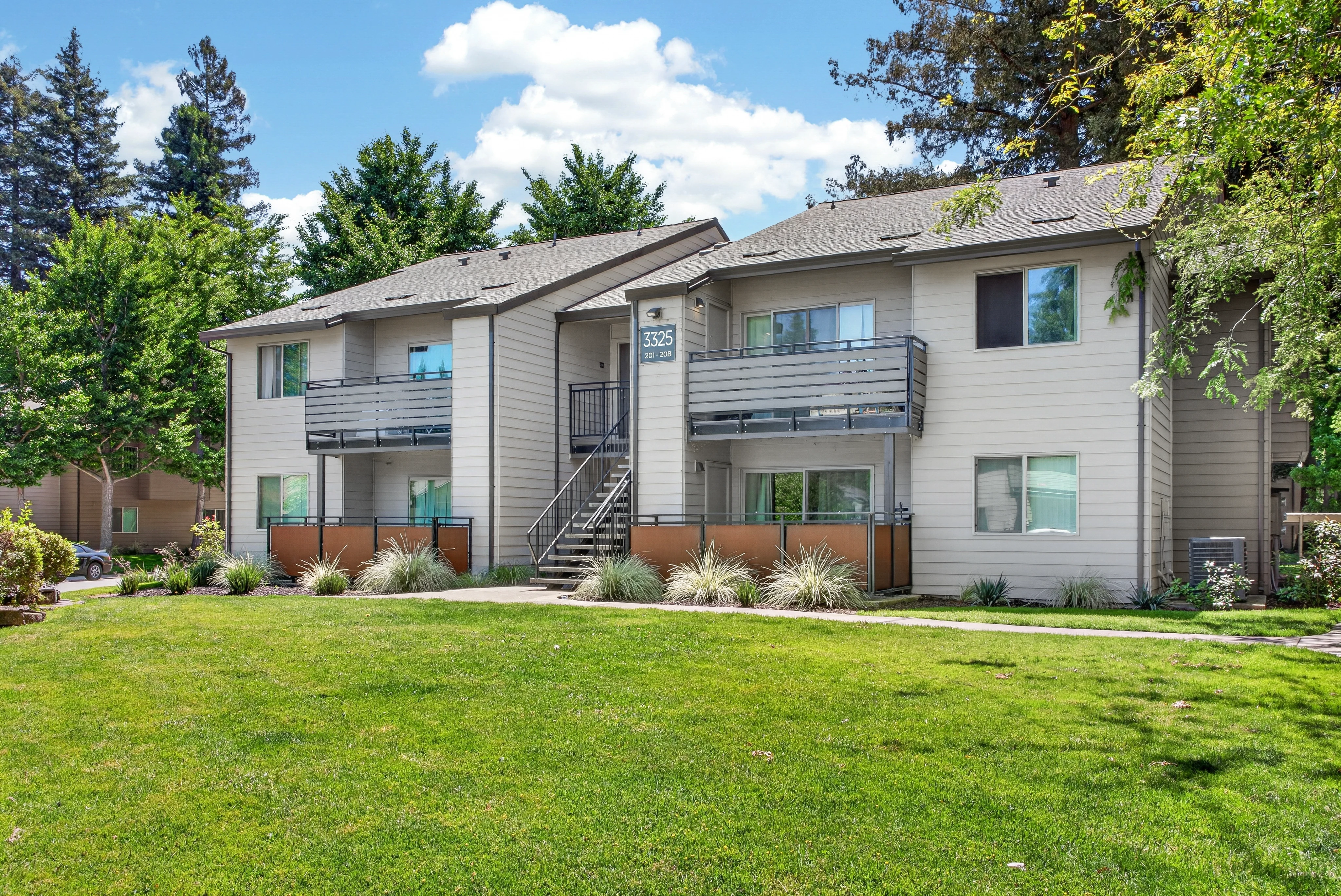 A two-story apartment complex with balconies, surrounded by a well-maintained lawn and trees under a clear blue sky.