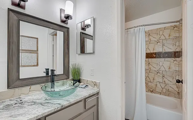 A modern bathroom with a stylish vanity, round glass sink, and decorative mirrors. The bathtub is enclosed with a white curtain, and the wall features tiled accents.