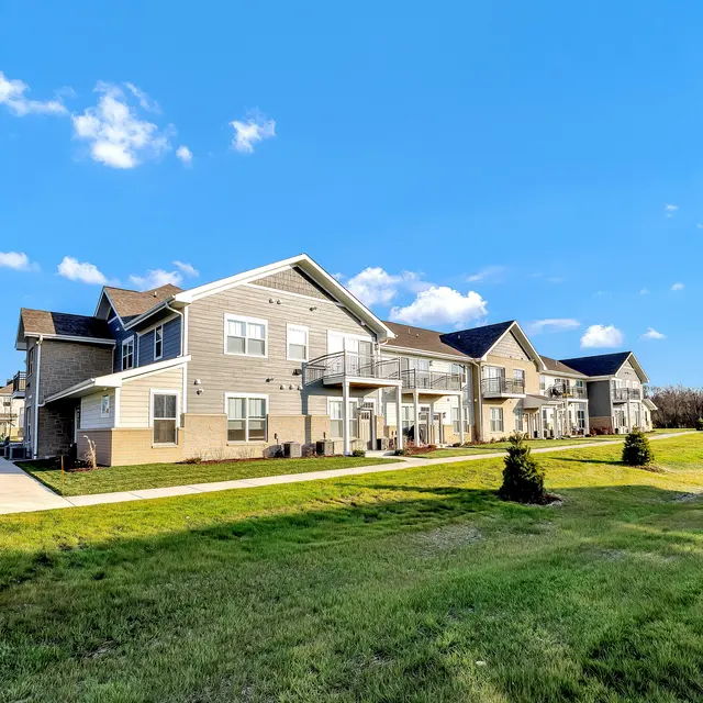A view of an apartment complex with multiple buildings set against a clear blue sky. The lawn is green with neatly manicured shrubs and a paved walkway.