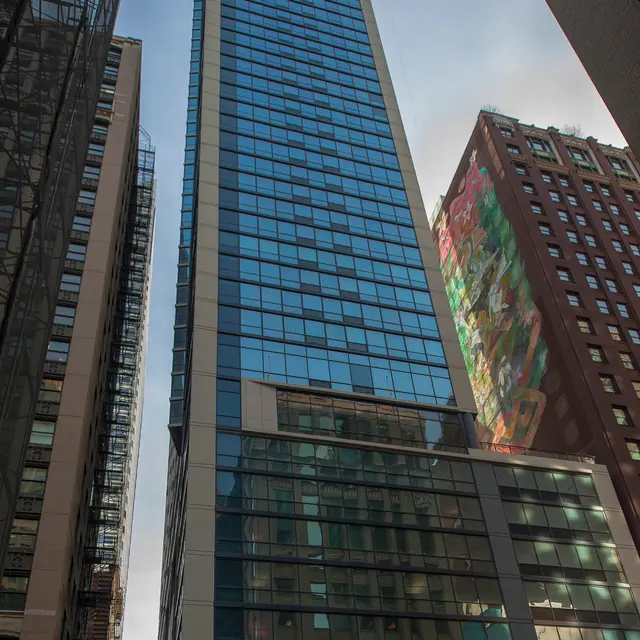 A tall glass skyscraper flanked by shorter buildings, with reflective windows and a colorful mural on one side.