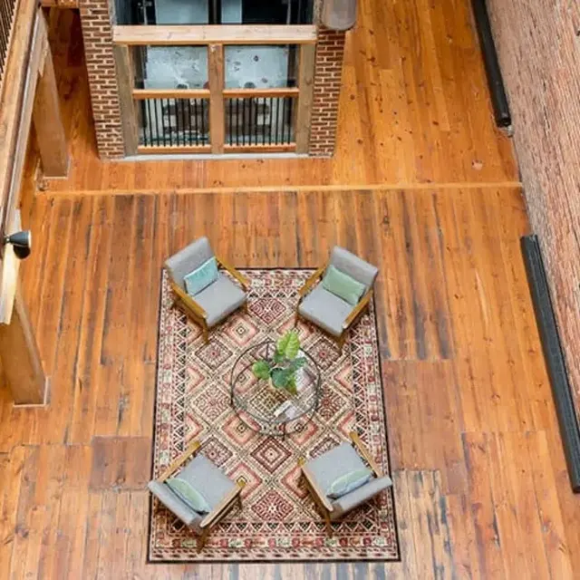 An overhead view of a cozy living space featuring a patterned rug, four gray armchairs, and a table with a plant in the center, surrounded by brick walls and wooden flooring.