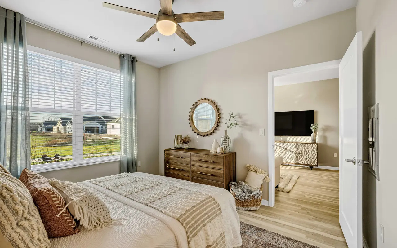 A cozy modern bedroom featuring a bed with decorative pillows, a wooden dresser, a circular mirror, and large windows providing natural light.