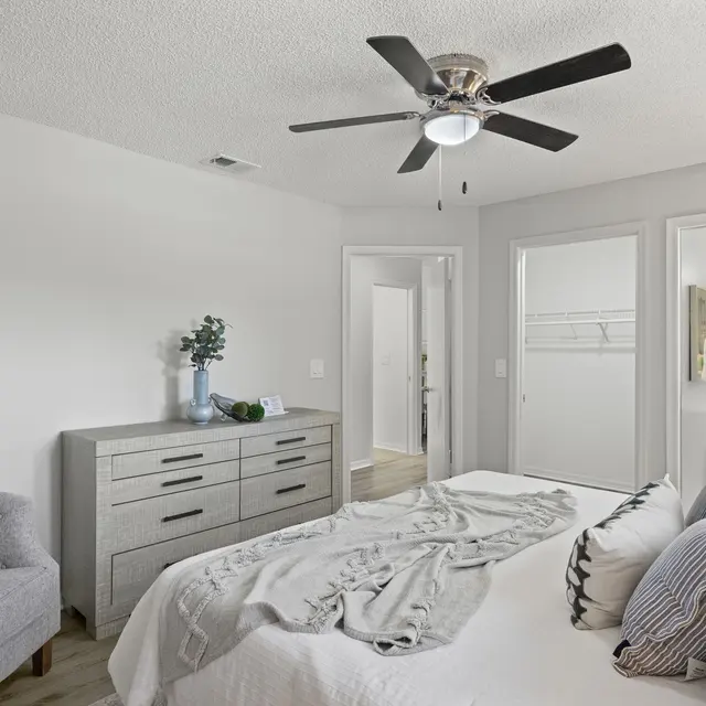 A modern bedroom featuring a light grey theme with a bed covered in soft bedding, a stylish dresser, and a grey couch with decorative pillows. The room has a ceiling fan and visible doors leading to an adjoining bathroom.