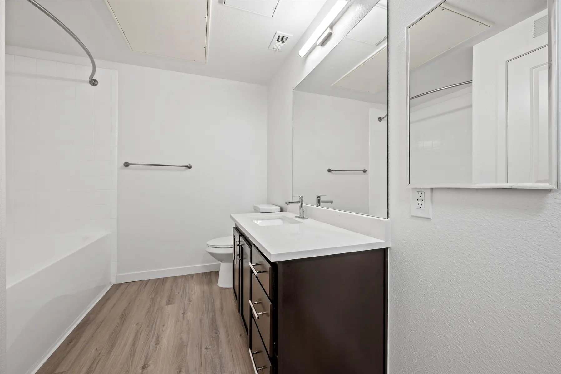 A modern bathroom featuring a dark wood vanity with a white countertop, a toilet, and a bathtub with a shower rod. The walls are painted white and the flooring is wood-like.