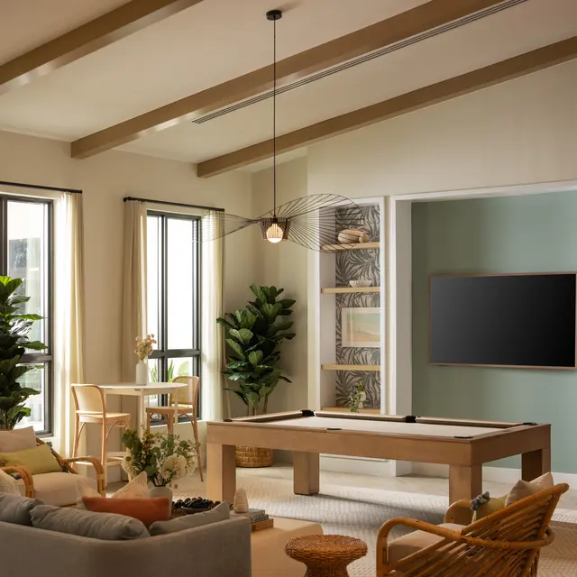 A spacious modern living room featuring a large pool table in the center, surrounded by a comfortable sofa, chairs, and decorative plants. Large windows allow natural light to fill the room, highlighting the stylish decor and layout.