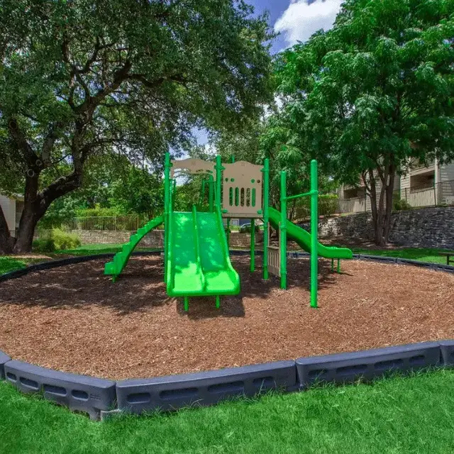 A vibrant playground with green slides and climbing structures surrounded by mulch and grass under a blue sky with clouds.