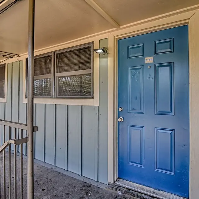 Exterior view of an apartment with a blue door, light blue wooden siding, and a covered entrance.