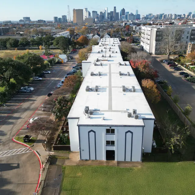 Aerial view of a multi-story apartment complex with flat roofs and a parking lot, featuring trees and a city skyline in the distance.