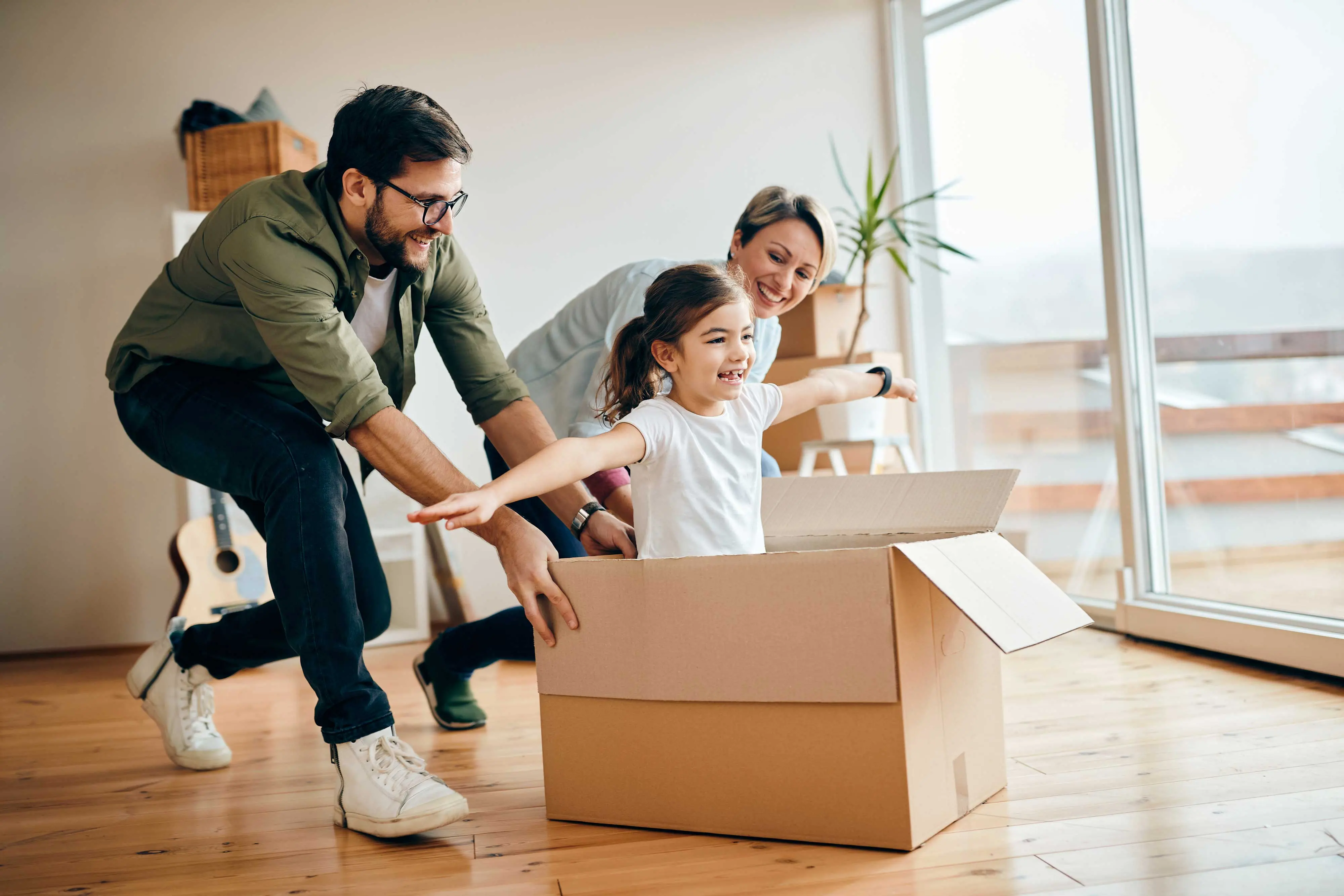 Family Playtime with Cardboard Box A family enjoying playtime in a bright room. A child is seated in a cardboard box with arms outstretched, while two adults push the box playfully. Large windows provide natural light and there are plants and a guitar in the background.