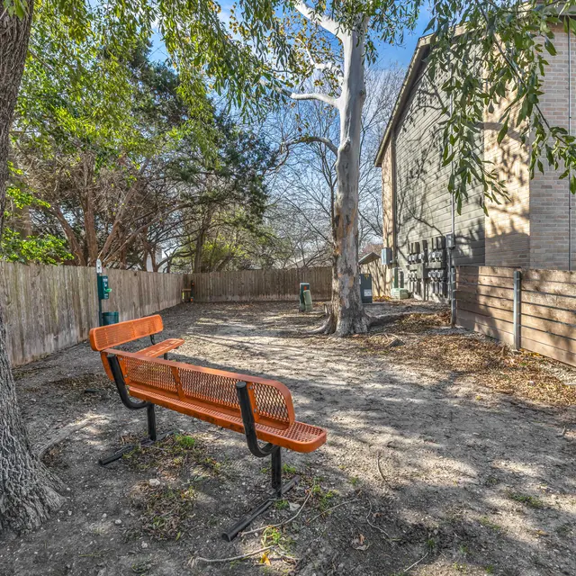 A fenced-in dog park with two benches, a pet relief station, and two large trees near The Enclave's apartment buildings.