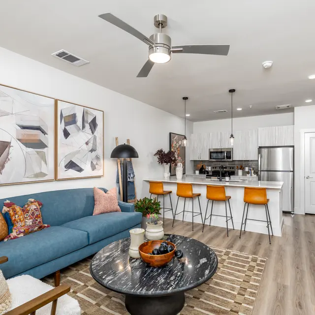 A modern living room featuring a blue sofa with colorful cushions, a round black coffee table, abstract wall art, and an open kitchen area with bar stools.