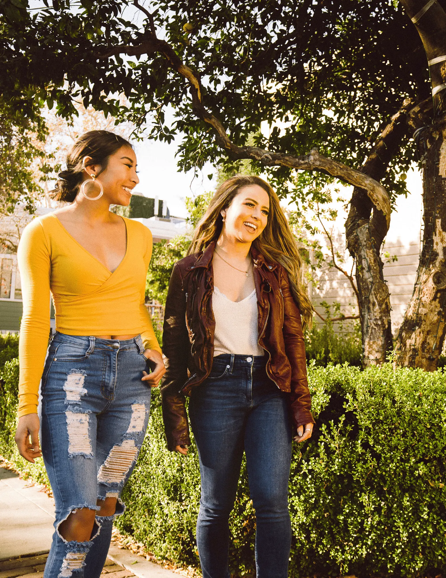 Two women wearing stylish outfits walking together in a sunny outdoor setting, smiling and laughing.