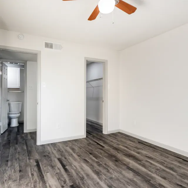 An empty room featuring a ceiling fan, wooden flooring, and a doorway leading to a bathroom.