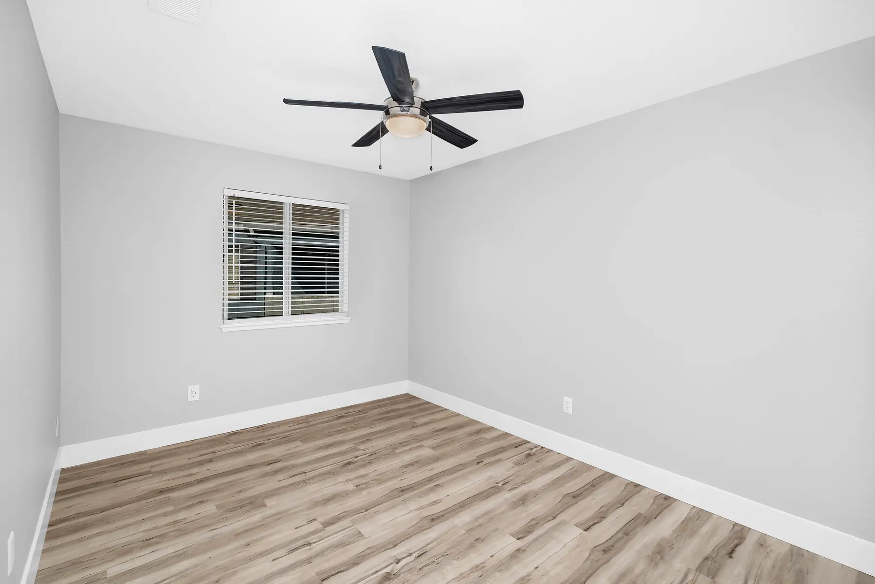 Empty room featuring a light gray wall and wooden floor, with a ceiling fan and a window with blinds.