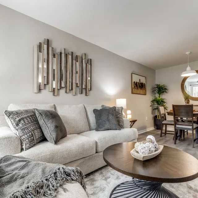 A modern living room featuring a light gray sofa, a round coffee table, and a dining area visible in the background. Decorative elements include a textured throw, plants, and framed wall art.