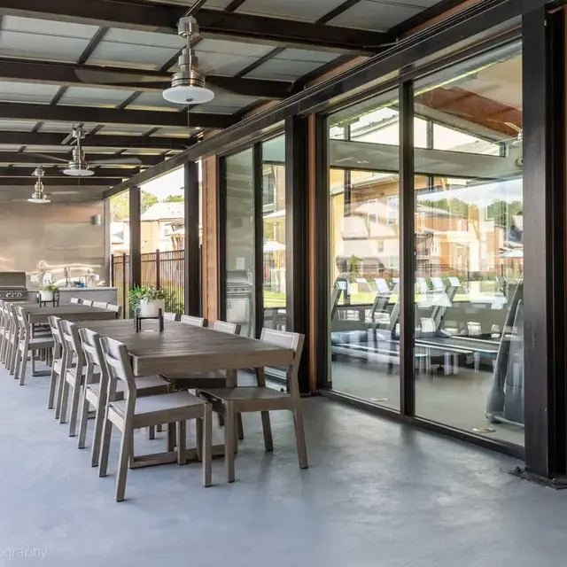 A modern outdoor dining area with a long wooden table and chairs, surrounded by large glass windows and an outdoor grill.