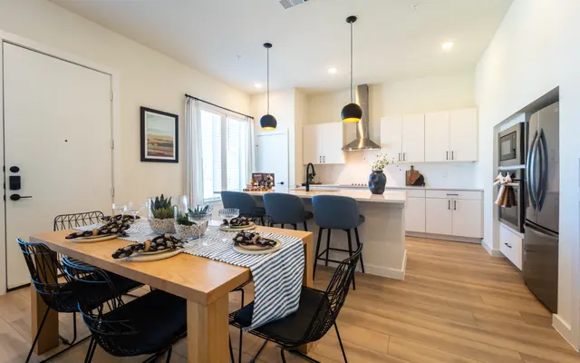 A modern kitchen and dining area featuring a wooden dining table with black wire chairs, set for a meal. The kitchen has white cabinetry, stainless steel appliances, and pendant lighting. Natural light enters through large windows, illuminating the space.