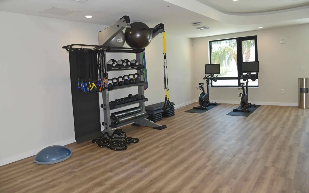 A modern gym interior featuring various workout equipment, including free weights, resistance bands, a stability ball, and two exercise bikes in a spacious room with wooden flooring and large windows.