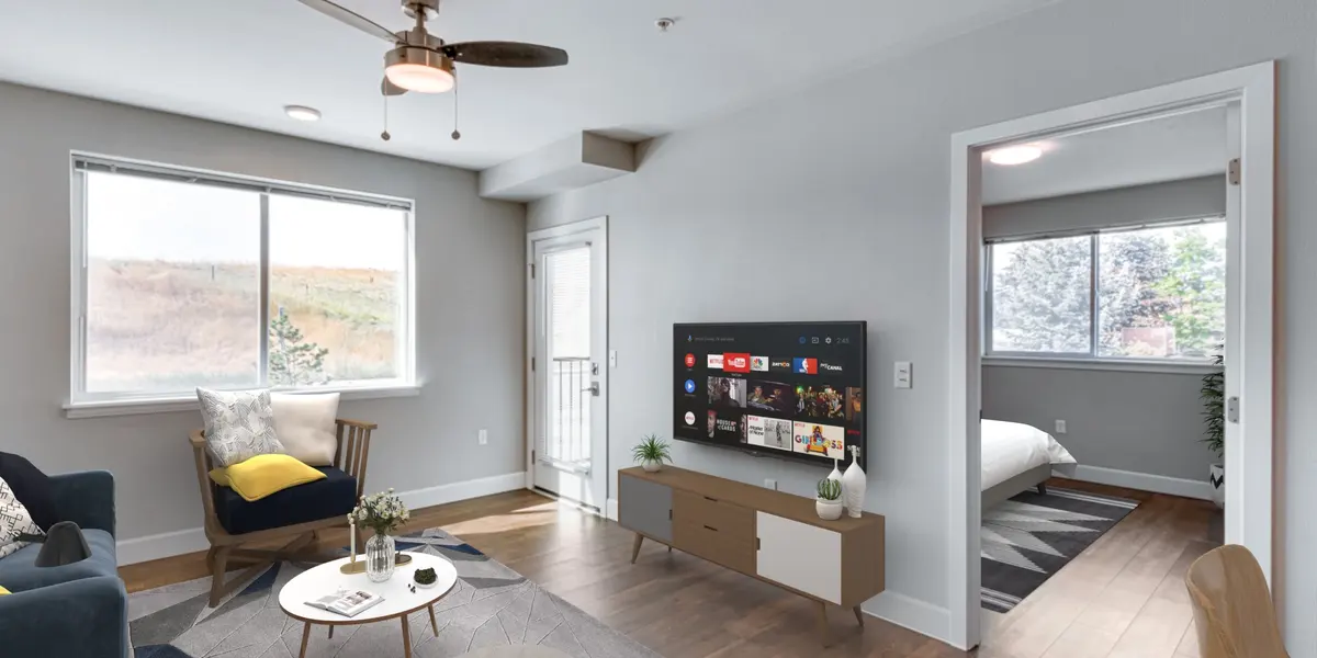 A modern living room featuring a mounted smart TV, a comfortable chair with a yellow pillow, and a coffee table with decor items. The room has large windows, wood flooring, and an open doorway leading to a bedroom with natural light.