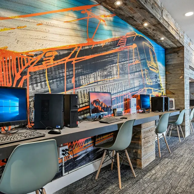 Modern Community Computer Area A modern computer area featuring multiple workstations, each with a desktop PC and stylish chairs. The wall behind is decorated with vibrant artistic designs.
