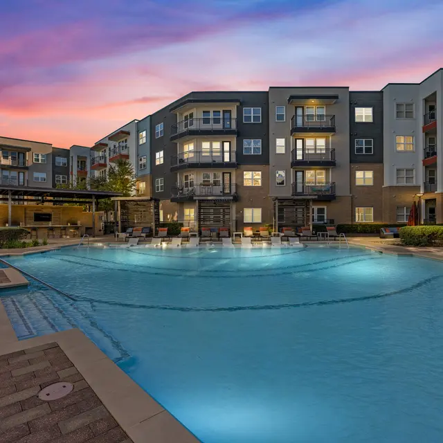 An apartment complex with a swimming pool at dusk, illuminated by soft lights, surrounded by greenery and outdoor seating.