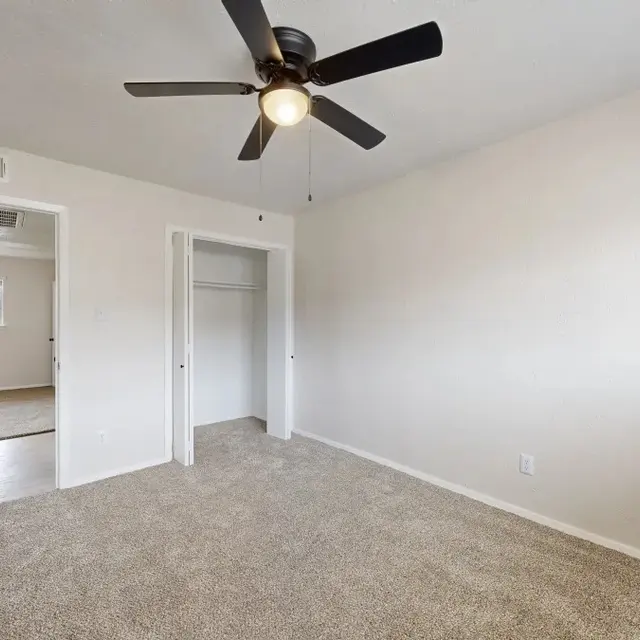 An empty bedroom with beige carpet, a ceiling fan, and two doorways leading to other rooms. The walls are light-colored and the room is well-lit.