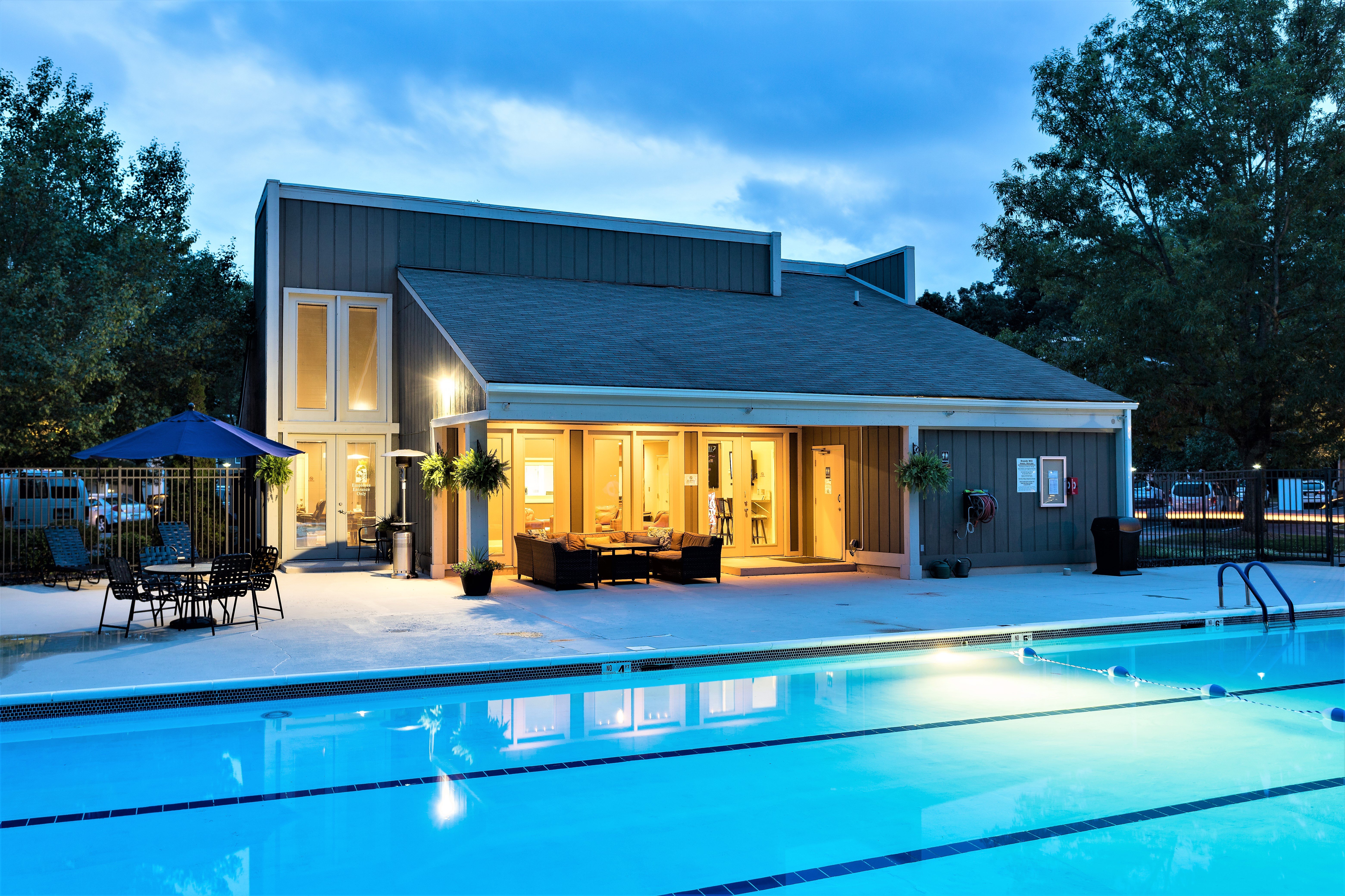 A serene swimming pool area during twilight with a modern building in the background. The pool is illuminated with lights and surrounded by trees. Patio chairs and tables are set around the pool, creating a welcoming atmosphere.