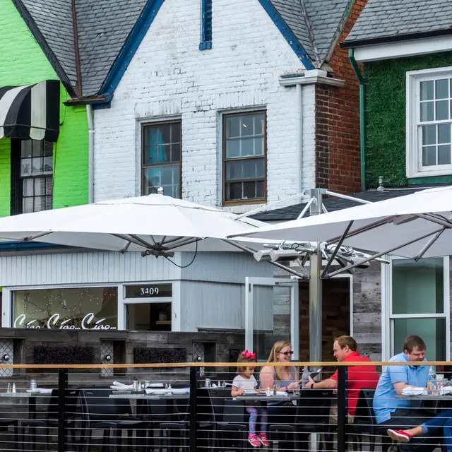 A lively outdoor dining area at a local Carytown, VA restaurant with people seated under large umbrellas. The background features colorful buildings with one having a bright green facade and another with a black and white striped awning.