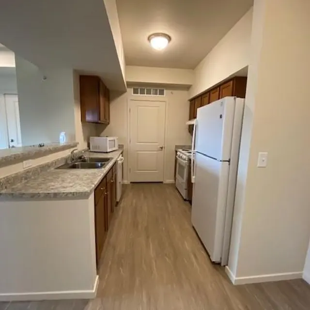A modern kitchen with wooden cabinets and granite countertops, featuring a sink, microwave, white refrigerator, and stove. The flooring is light wood, and there is ample natural light coming from the adjacent room.