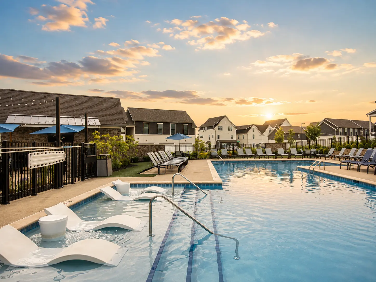A scenic view of a swimming pool area with lounge chairs, surrounded by houses, under a sunset sky.