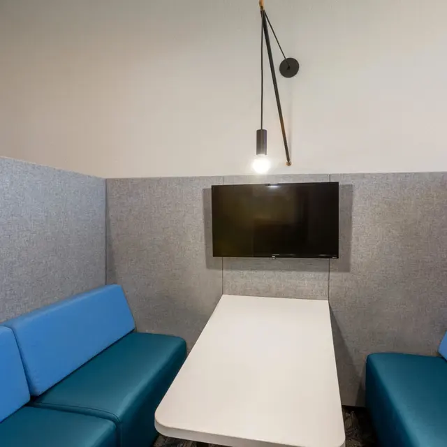 A cozy meeting area featuring blue cushioned benches and a white table, with a flat-screen TV mounted on a gray partition wall and a hanging light fixture.
