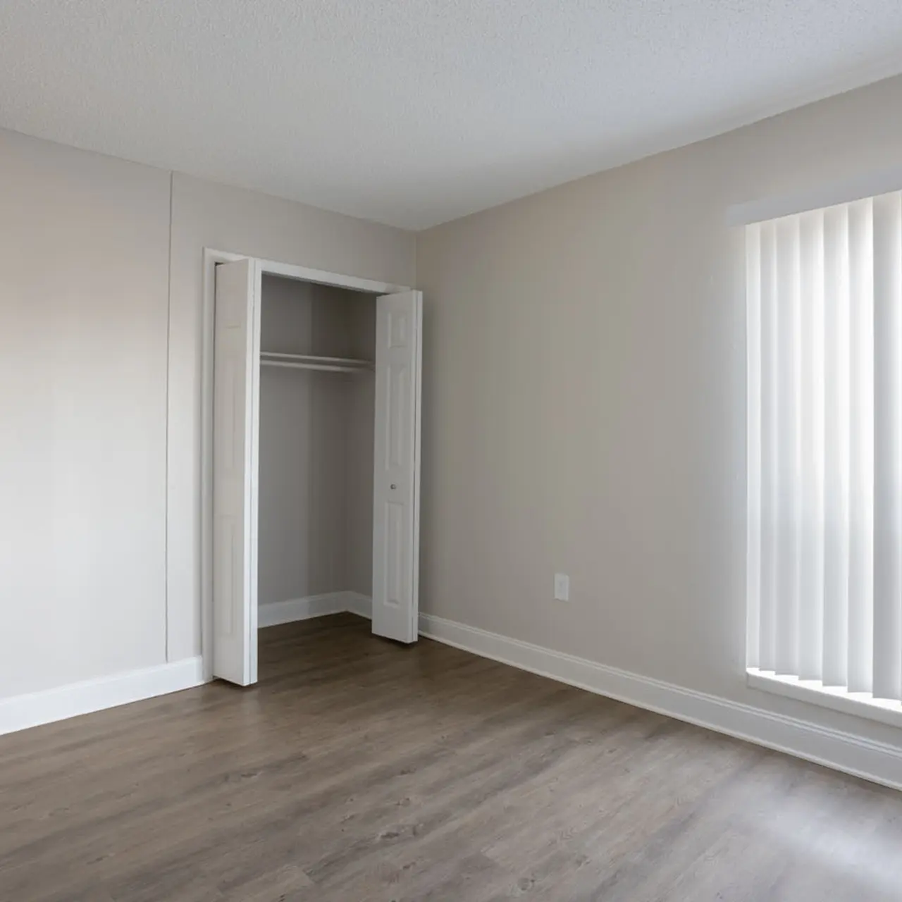 A bright, empty room with light grey walls, wooden flooring, and a closet. There is a window with vertical blinds allowing natural light to enter, and a closed door on the left.