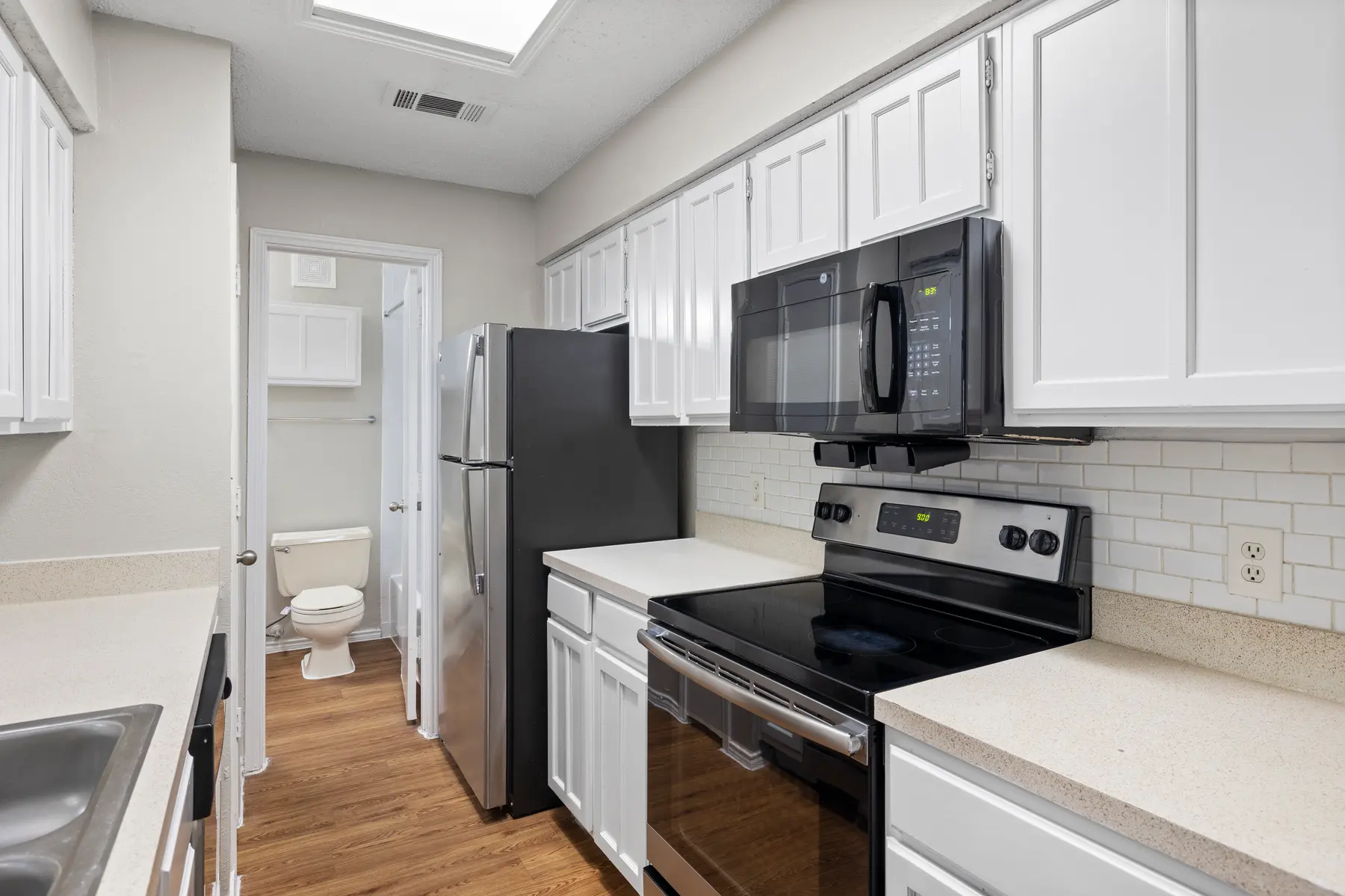 A modern kitchen featuring white cabinets, stainless steel appliances including a microwave and stovetop, with a light-colored countertop and an open layout. A glimpse of a bathroom is visible through an open door.