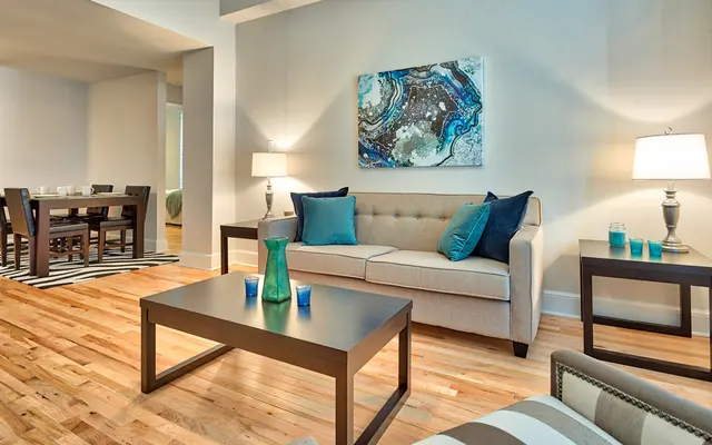 Modern Living Room Decor A modern living room featuring a light-colored sofa with blue and teal cushions, a wooden coffee table, and a pair of lamps. The floor is hardwood, and there is a dining area visible in the background with a dark wooden dining table and chairs. A decorative wall art piece with blue and brown tones hangs above the sofa. The space is well-lit and stylishly decorated with a cozy feel.