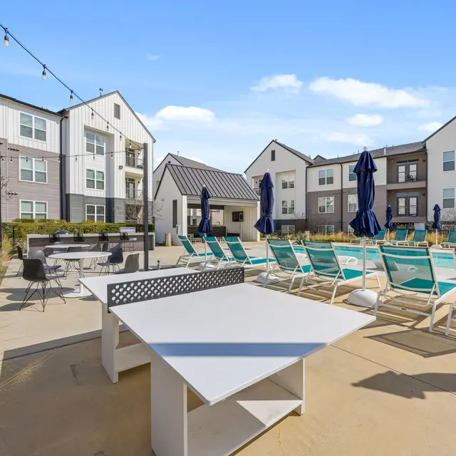 A pool area at an apartment complex featuring lounge chairs, umbrellas, a table tennis table, and landscaped surroundings.